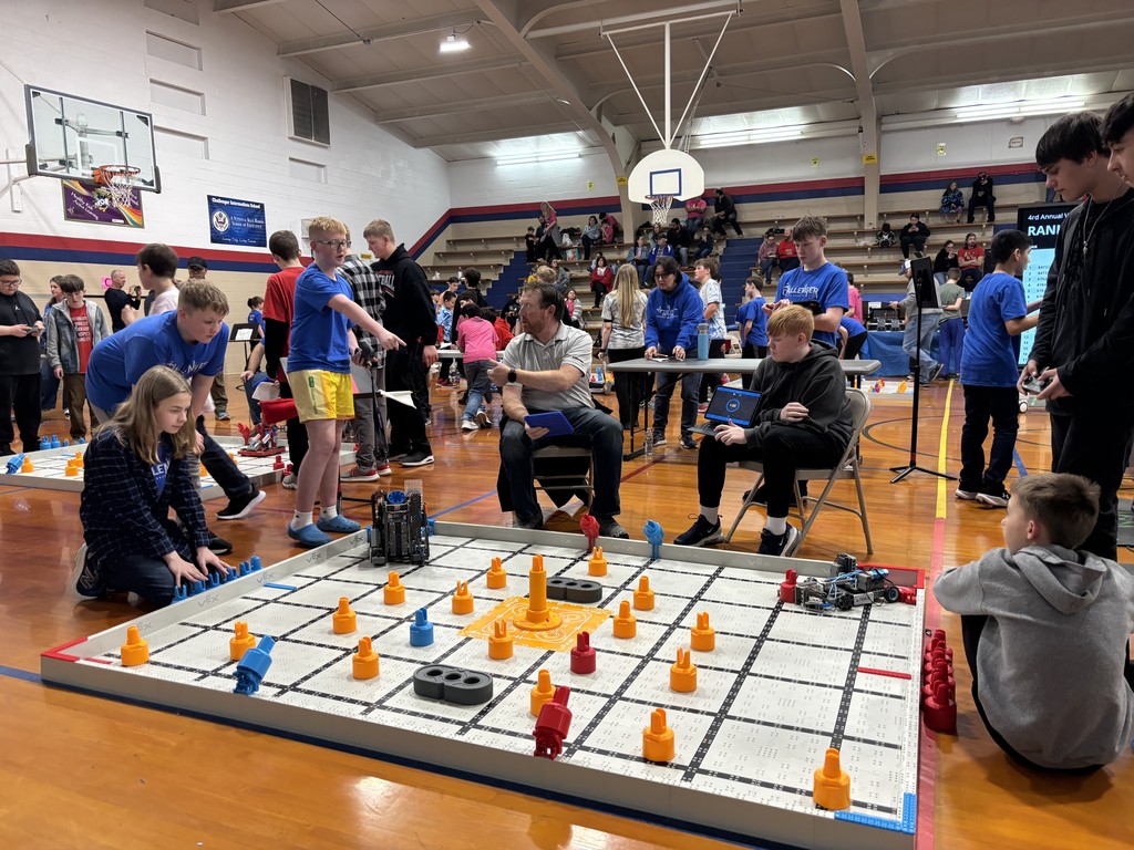 A group of students at a robotics tournament. on the floor is the robotics challenge board with various obstacles for their robots to work through. 