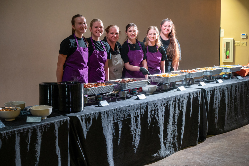 Six people wearing aprons stand smiling behind a buffet table filled with trays of food. The atmosphere is warm and inviting, suggesting a catered event.