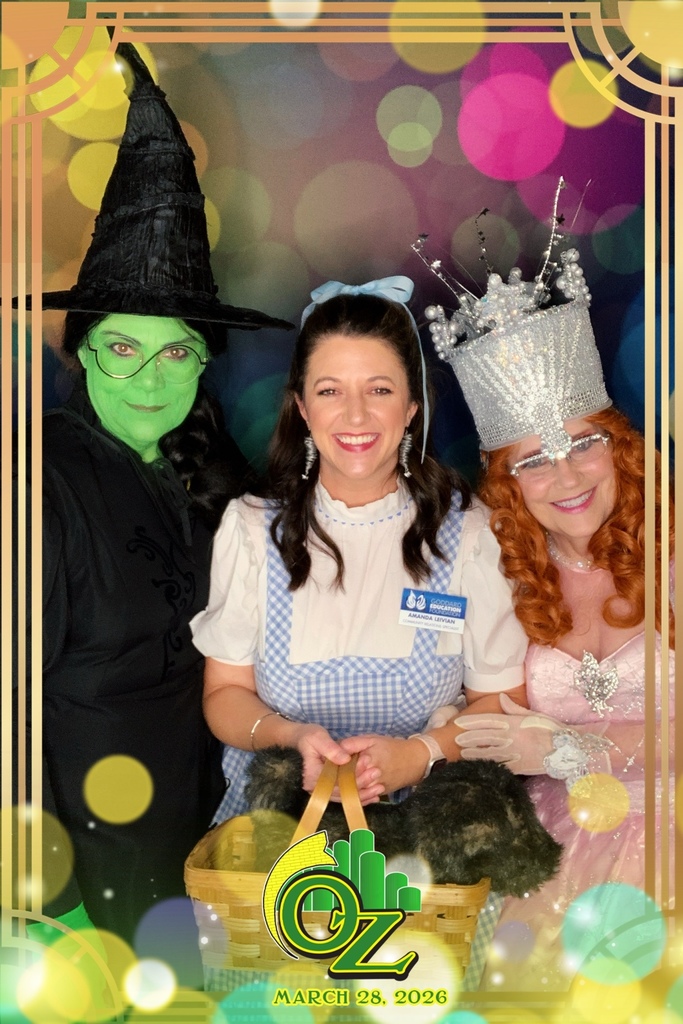 Three people in colorful costumes resembling characters from "The Wizard of Oz" pose together, smiling cheerfully. The date reads March 28, 2026.