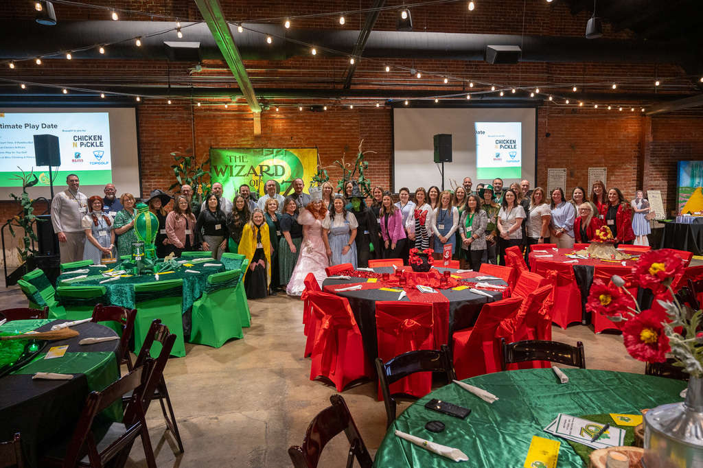 A large group of people pose smilingly in front of a "Wizard of Oz" backdrop. Tables with colorful red and green tablecloths fill the festive room.