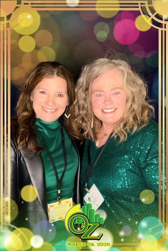 Two women smiling brightly in front of a colorful, bokeh background. They wear green tops and lanyards, framed with an "Oz" theme and date.