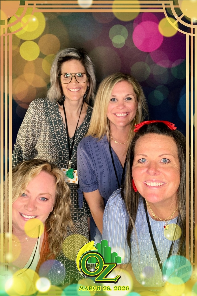 Four smiling women pose against a colorful, bokeh background. They wear lanyards and casual attire. A border and "OZ" with a date suggest a themed event.