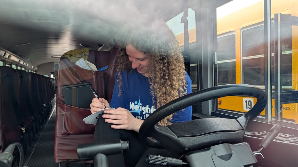 A person with curly hair, wearing a blue "Neighbors" shirt, writes on a notepad inside a school bus. They appear focused and content.