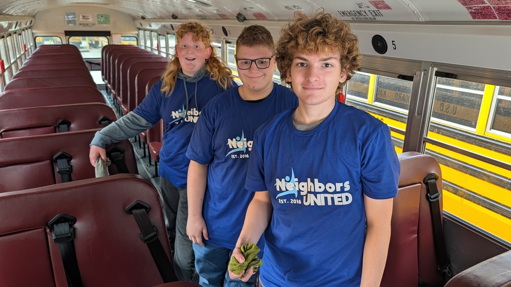 Three smiling teenagers wearing blue "Neighbors United" T-shirts stand inside a school bus. They convey a sense of teamwork and community spirit.