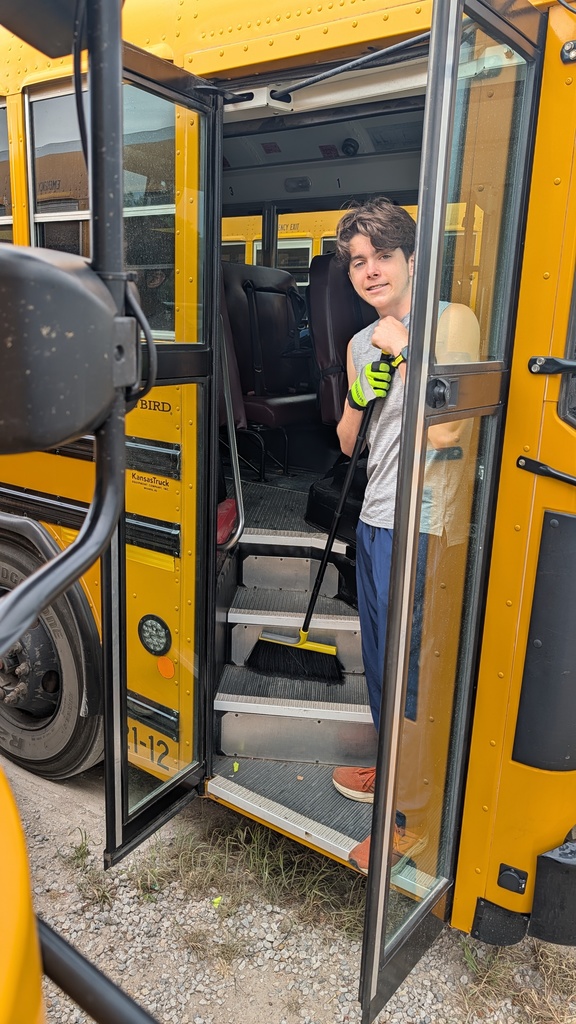A person stands in the doorway of a yellow school bus with a broom, wearing gloves and smiling. The scene conveys a sense of care and responsibility.