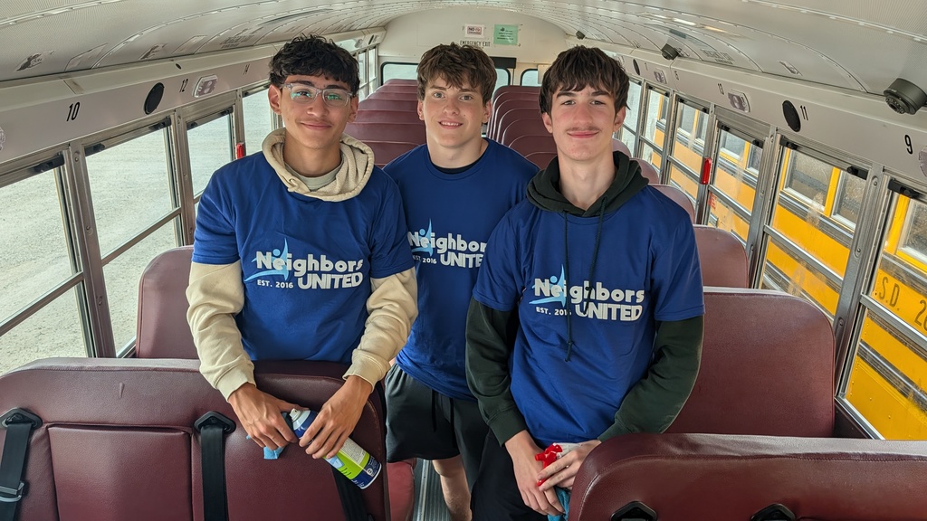 A group of three, smiling, teenaged boys, in blue "Neighbors United" shirts, clean a school bus, creating an energetic, positive community service atmosphere.