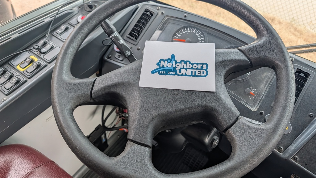The image shows a black steering wheel inside a vehicle. A white card with "Neighbors United Est. 2016" in blue text rests on the wheel, surrounded by control panels.