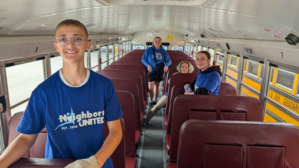 A group of smiling teens in blue "Neighbors United" shirts clean a school bus, creating an energetic, positive community service atmosphere.