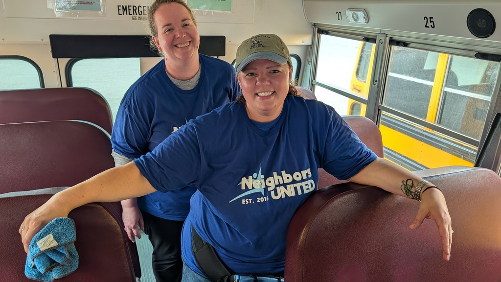 Two people smile inside a school bus, wearing blue "Neighbors United" T-shirts. One holds a cleaning cloth, suggesting teamwork and community spirit.