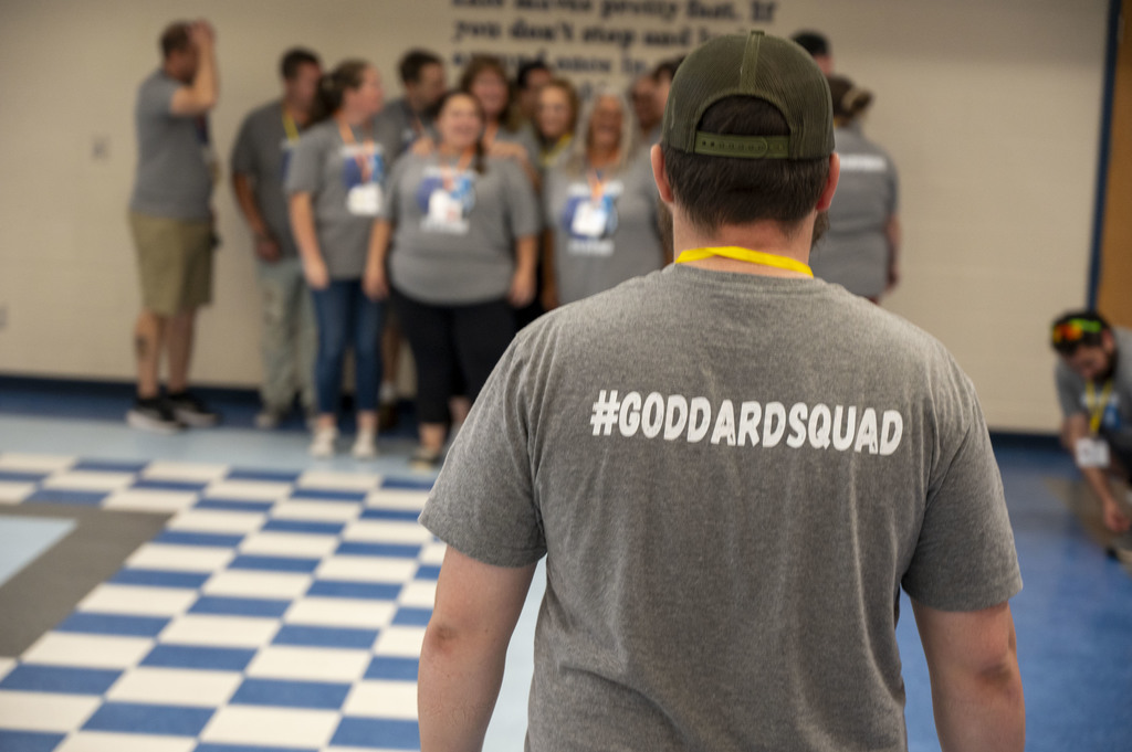 A group of people in gray shirts stand together, with one person in front wearing a "#GODDARDSQUAD" shirt. The floor has a blue and white checkered pattern.
