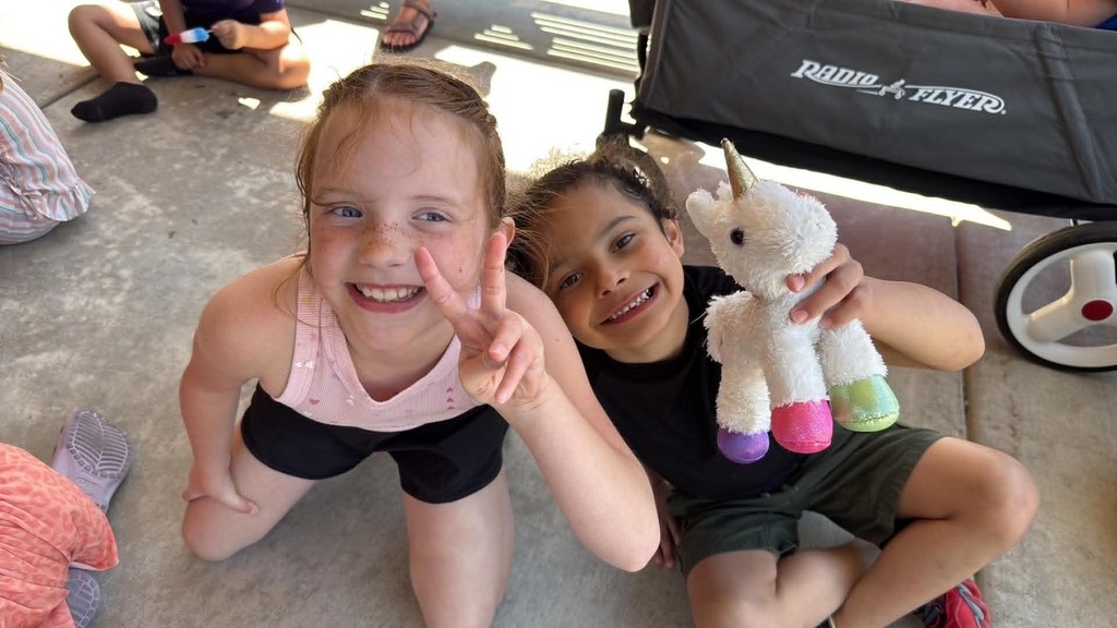 Two smiling children sitting on the ground. The one on the left flashes a peace sign, while the one on the right holds a plush unicorn. A wagon is in the background.