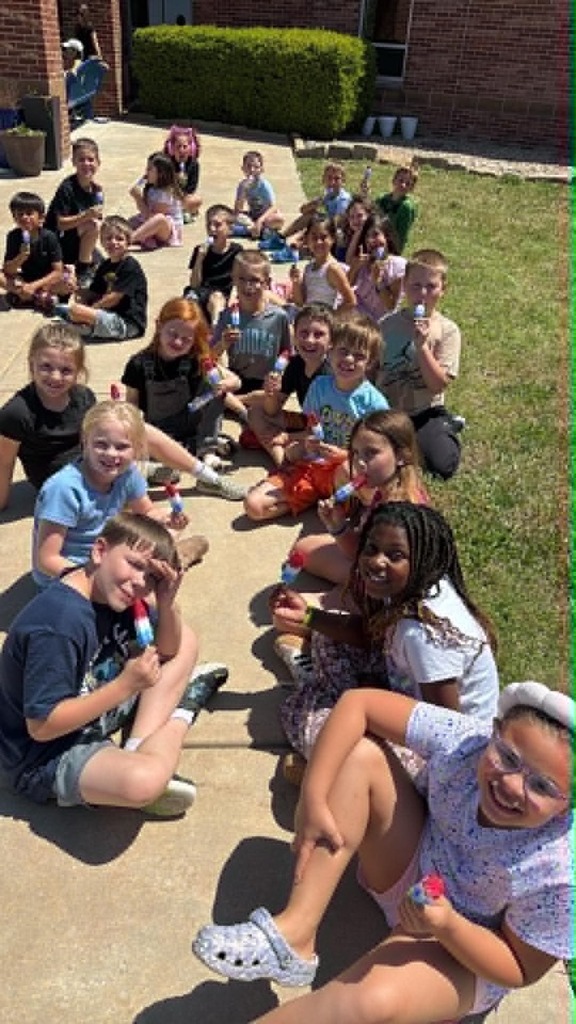 A group of children sit happily on a sunny walkway, enjoying colorful ice pops. Their faces are joyful, conveying a sense of fun and celebration.
