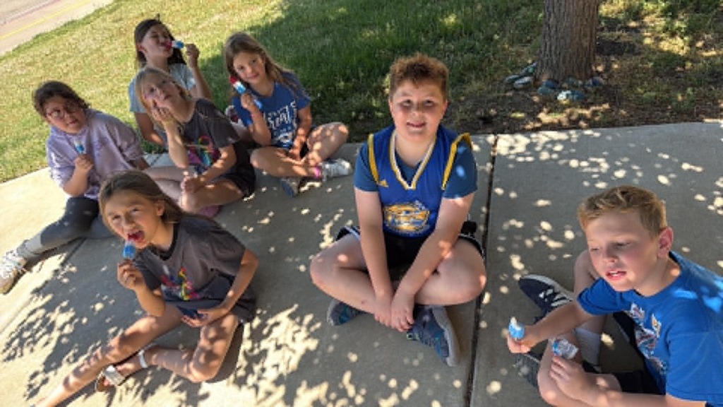 A group of children sits on a sunlit sidewalk under a tree, eating colorful ice pops. They appear happy and relaxed, enjoying a sunny day together.