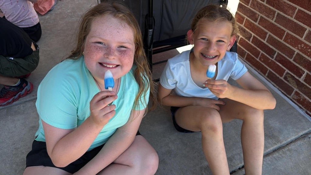 Two smiling children sit on a concrete surface enjoying blue popsicles. They appear joyful, sitting beside a brick wall with a relaxed, cheerful atmosphere.
