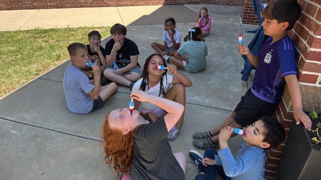 Ten children sit joyfully on a sunny patio, savoring colorful popsicles. Their expressions and relaxed poses convey a sense of enjoyment and camaraderie.