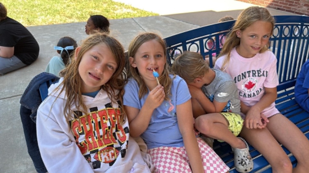 Four children sit on a bench smiling, with another child nearby looking down. It's a sunny day, and the atmosphere feels joyful and relaxed.