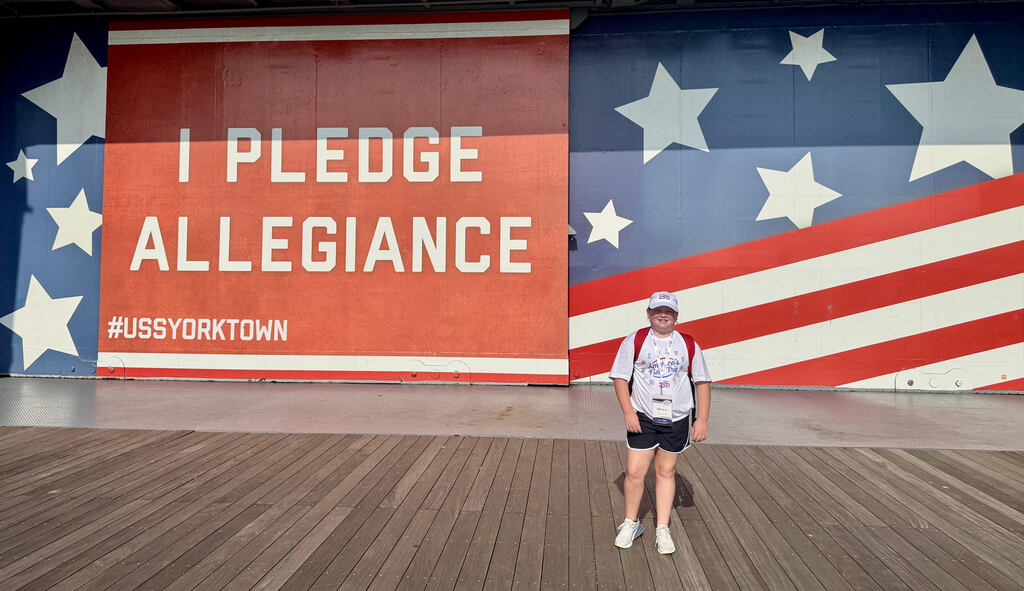 A 5th grade student stands in front of a large mural painted with red and white stripes on a blue background with white stars popping out. Off to the left is a giant red sign with the words "I pledge Allegiance #ussyorktown" written on it. 
