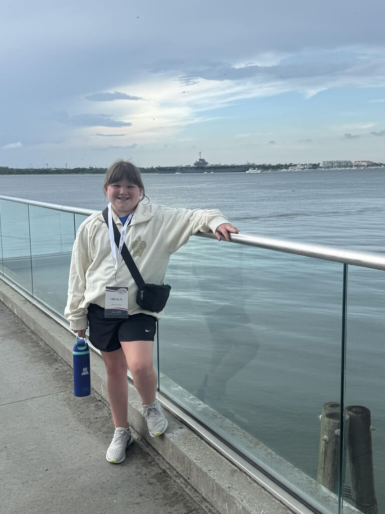A 5th grade girl stands on a cement pier way leaning against a glass railing with the view of the ocean behind her, and several military ships in the distance