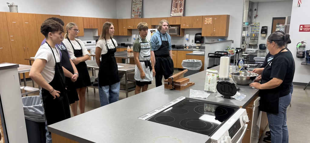 background: culinary classroom with students in aprons and 2 lunch ladies demonstrating at the kitchen counter