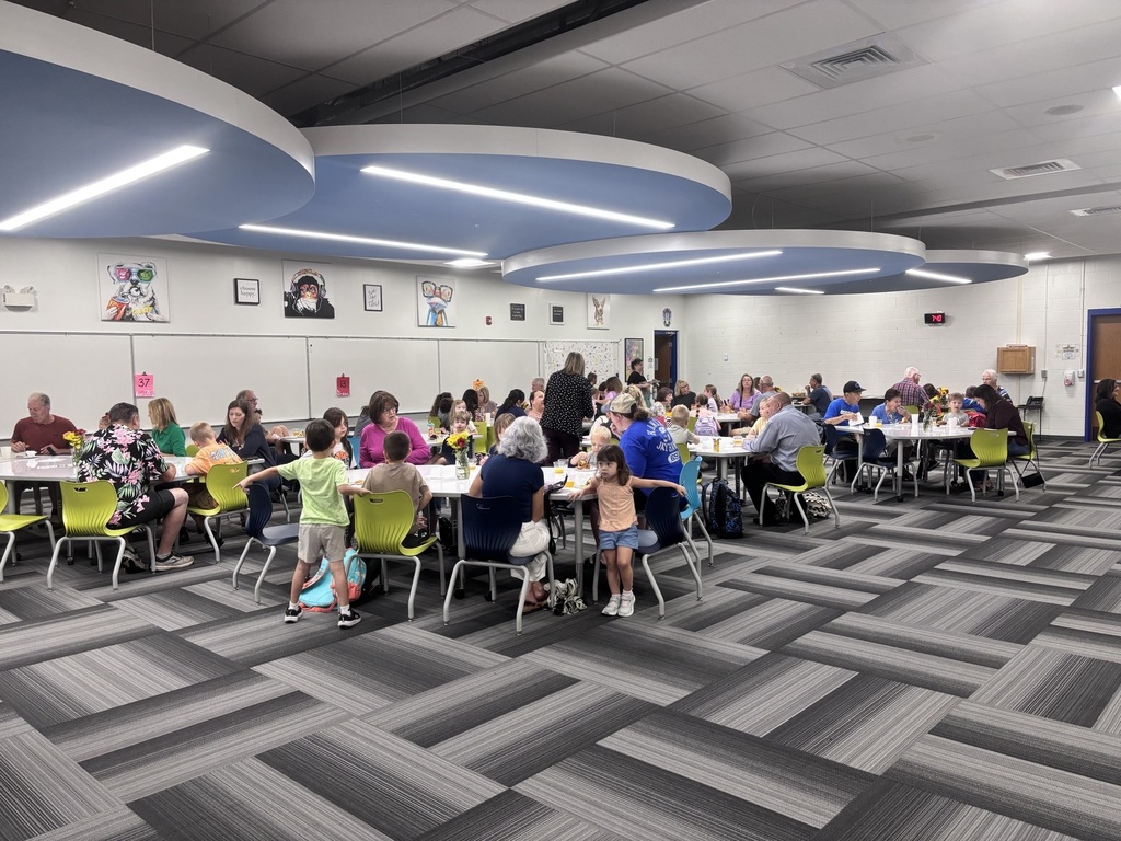 A lively school cafeteria filled with children and adults eating together, colorful chairs, Halloween costumes, and bright modern ceiling lights.