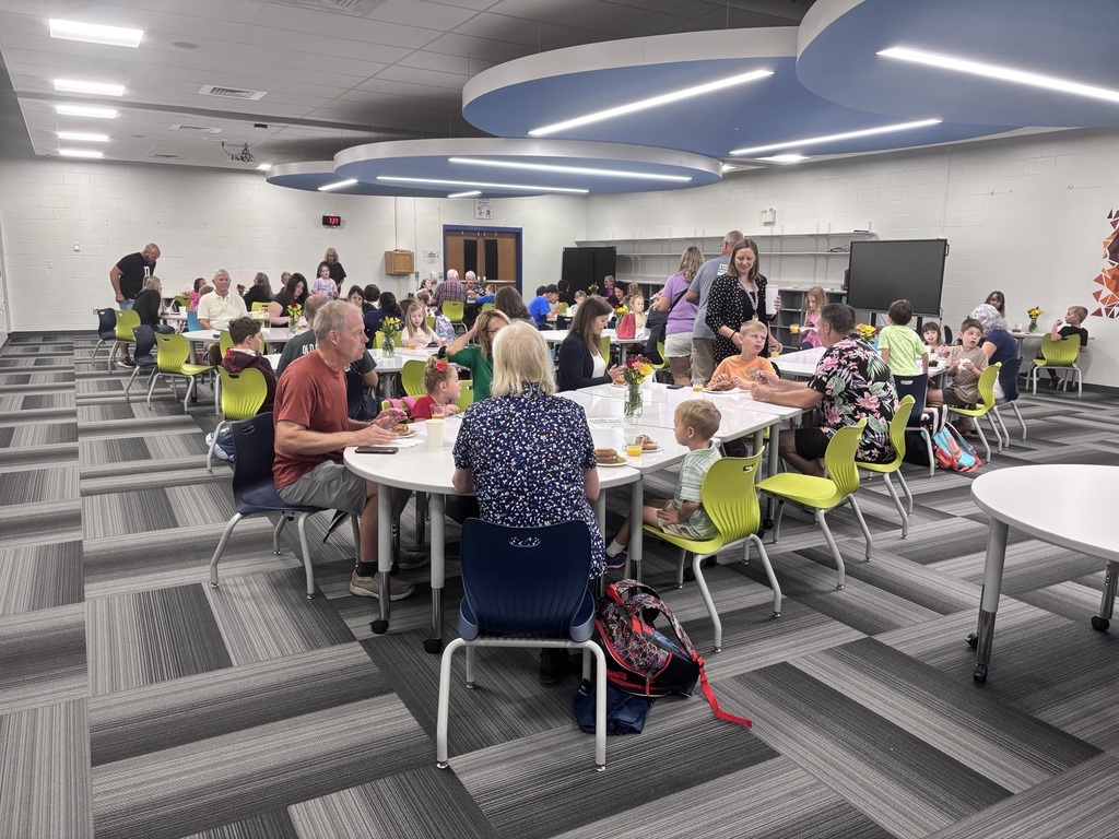A lively school cafeteria filled with children and adults eating together, colorful chairs, Halloween costumes, and bright modern ceiling lights.