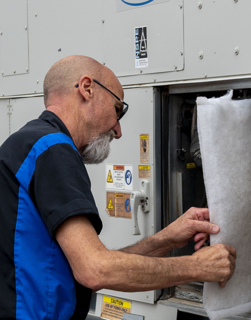 A male technician in blue-and-black workwear inspects industrial equipment, carefully lifting a white filter curtain beside safety labels; focused, hands-on work.