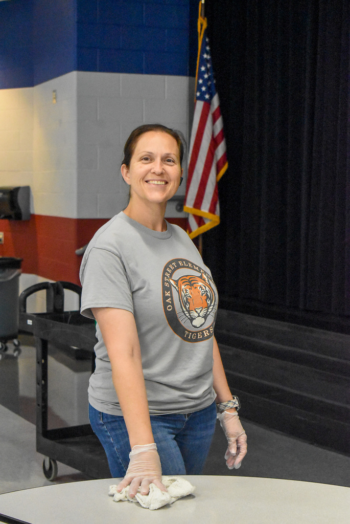 A smiling woman in a gray tiger-logo T-shirt wipes a table in a school gym, with an American flag and stage curtain behind her; the mood is warm and helpful.