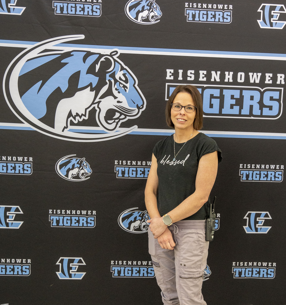 A smiling woman stands before an Eisenhower Tigers backdrop featuring blue-and-white tiger logos and team lettering, in a proud, welcoming portrait.