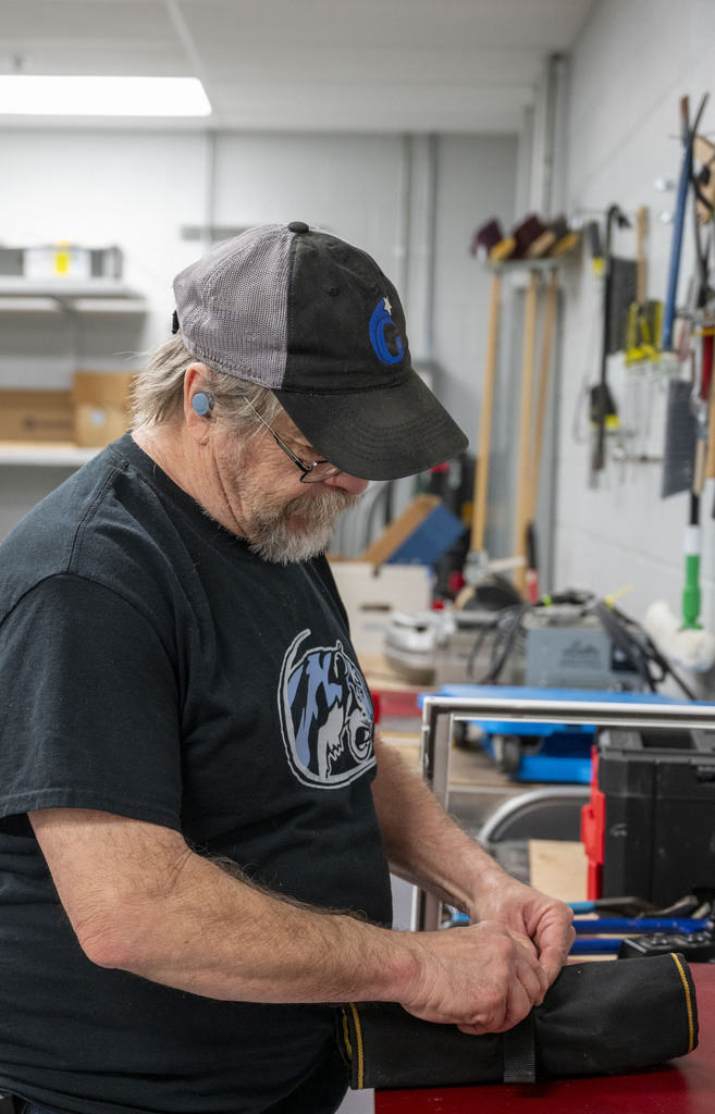 A gray-bearded man in a baseball cap concentrates on adjusting a black protective glove in a bright, tool-filled workshop.