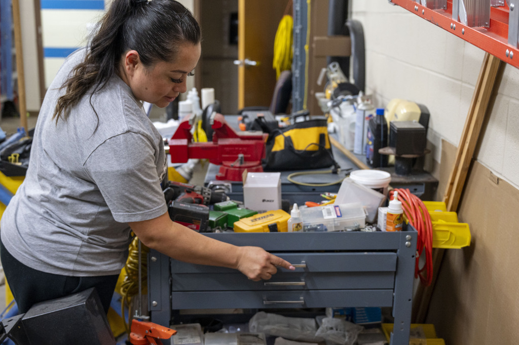 A woman in a gray shirt searches a crowded workshop tool cart, surrounded by tools and supplies; the focused scene conveys hands-on work.