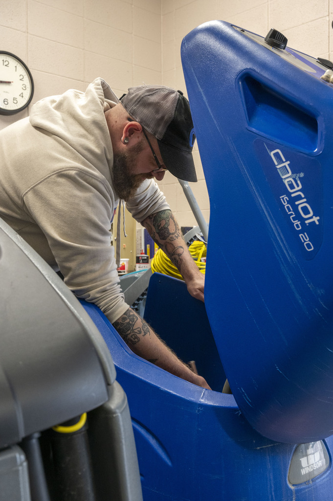 A tattooed maintenance worker in a cap leans into an open blue floor scrubber, inspecting its tank in a tiled room; focused, hands-on work.