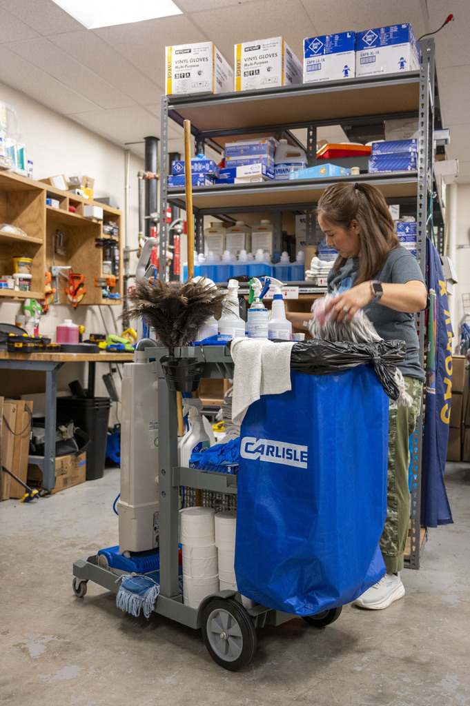 A woman cleans a room with a wheeled supply cart, blue Carlisle laundry bag, mop, and stocked shelves in a busy janitorial closet.