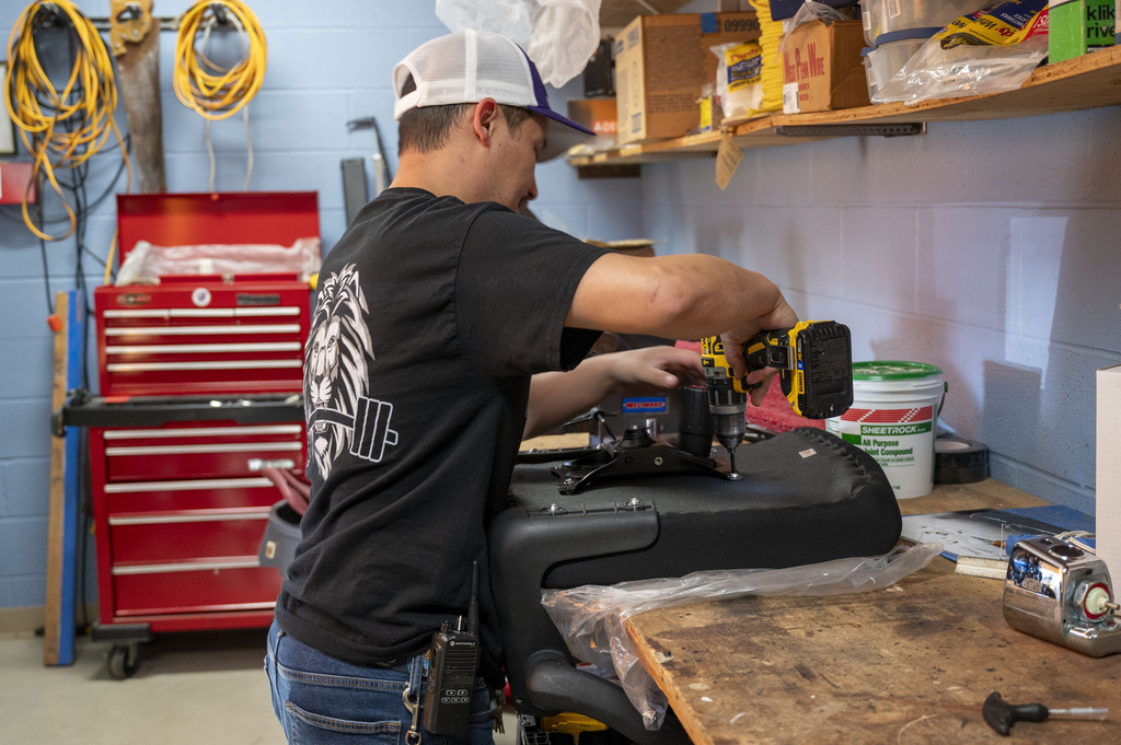 A focused repair technician drills into a padded black seat at a cluttered workshop bench, with red tool drawers, shelves, and supplies nearby.