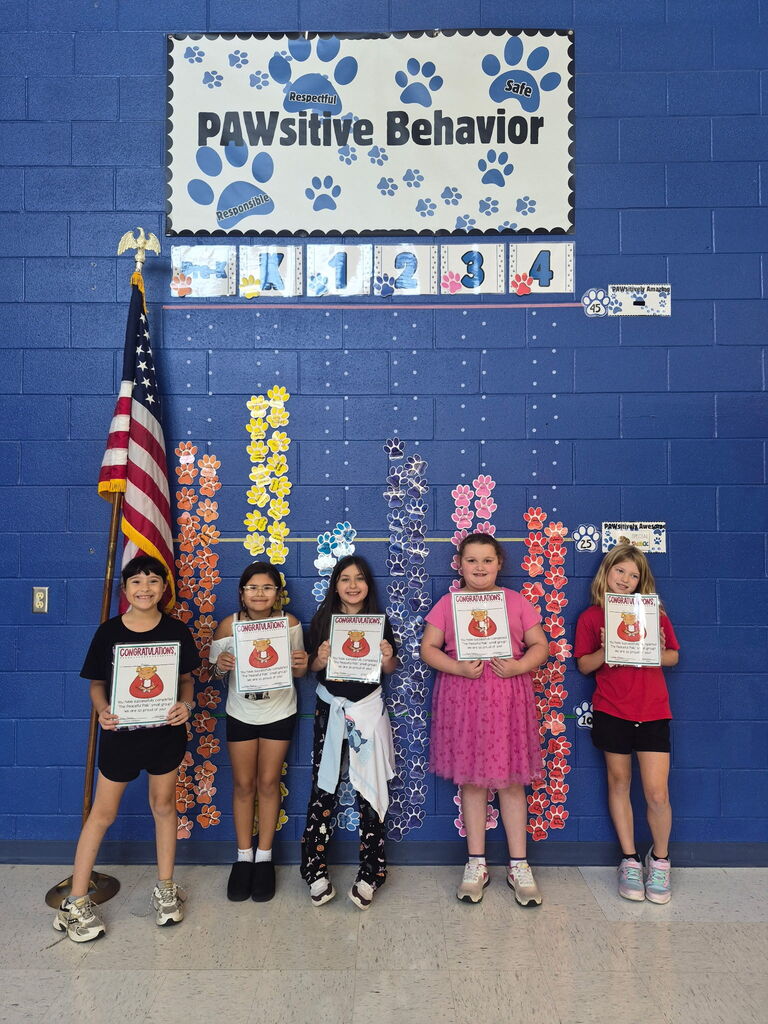 Four smiling children hold certificates beside colorful paw-print behavior charts on a blue classroom wall beneath a “PAWSitive Behavior” sign, celebrating achievement.