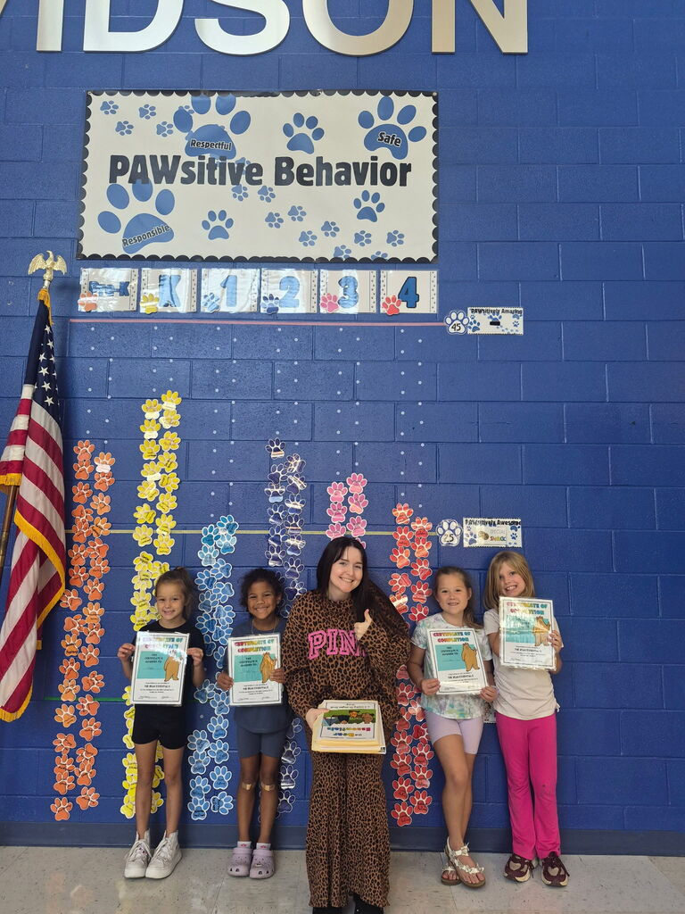 Four smiling children hold certificates beside colorful paw-print behavior charts on a blue classroom wall beneath a “PAWSitive Behavior” sign, celebrating achievement.