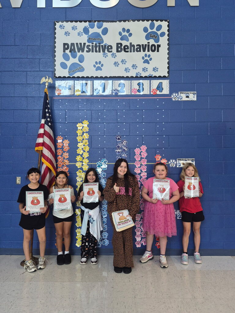 Four smiling children hold certificates beside colorful paw-print behavior charts on a blue classroom wall beneath a “PAWSitive Behavior” sign, celebrating achievement.