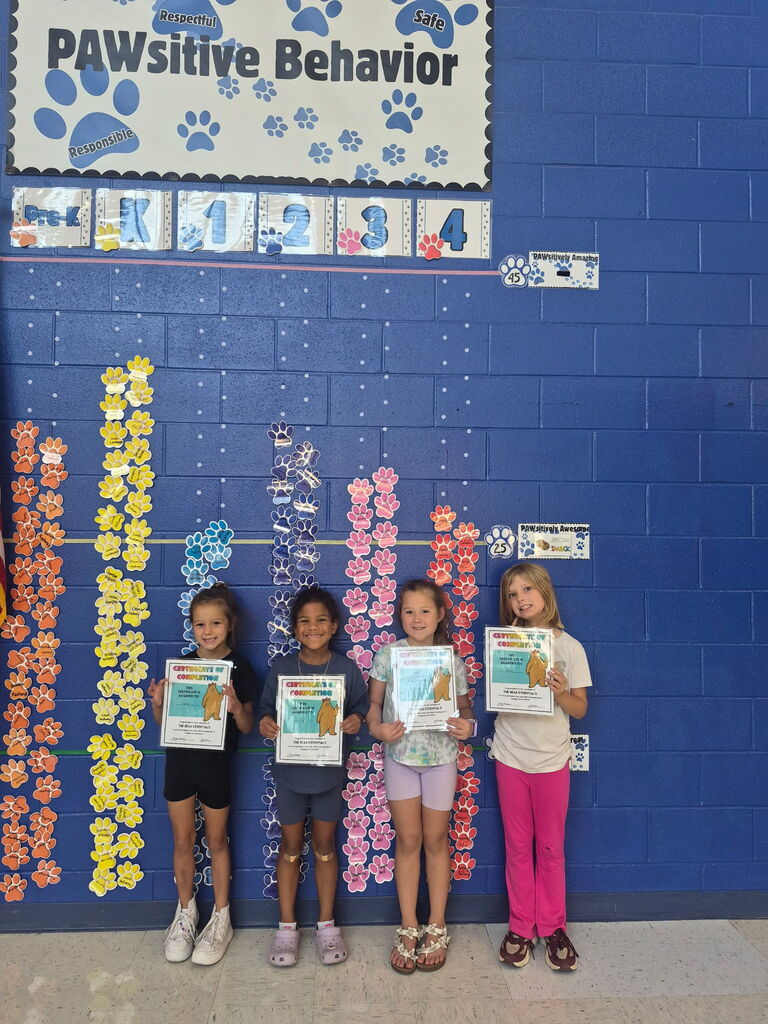 Four smiling children hold certificates beside colorful paw-print behavior charts on a blue classroom wall beneath a “PAWSitive Behavior” sign, celebrating achievement.