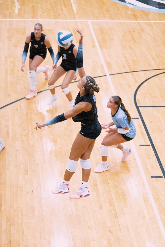 background: gym floor volleyball court, 4 IKE girls v-ball players dressed in black & blue, 3 with knees bent squatting, 1 jumping up to spike the ball.