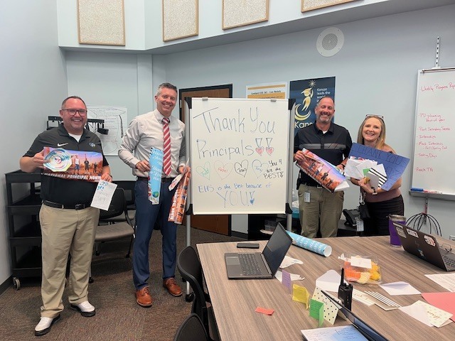 background: office conference room with 4 principals standing by a table with cards inhand and a white board message; text: Thank you principals, you are the best! EHS is the best because of you!