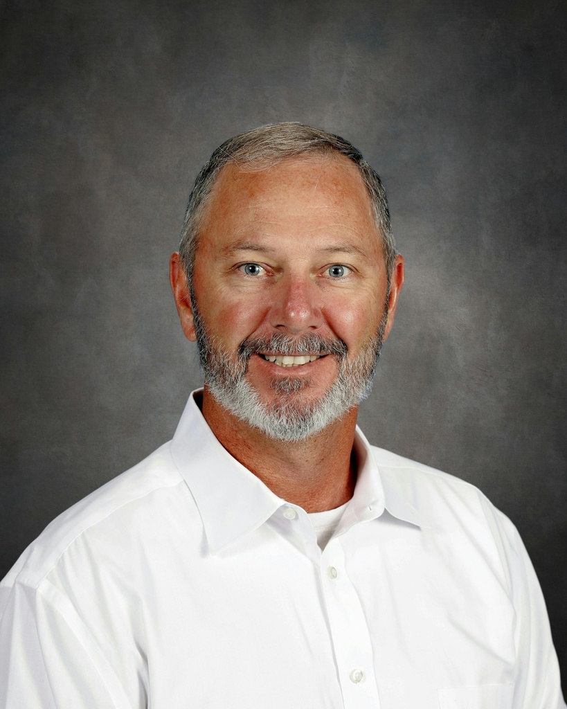 Studio portrait of a man with short graying hair and a salt-and-pepper beard, smiling warmly in a white collared shirt against a gray backdrop.