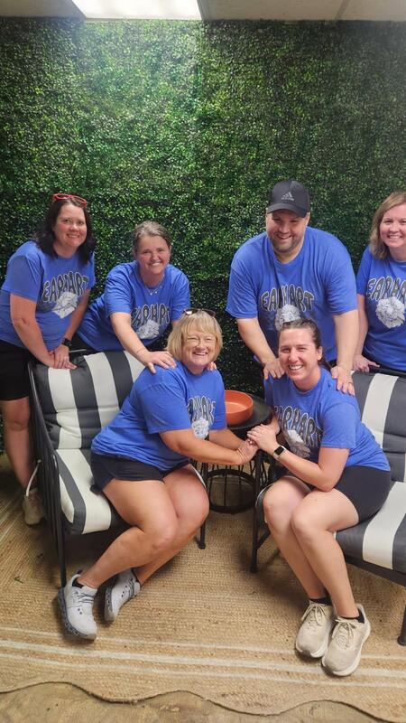 A group of six individuals, wearing matching blue t-shirts, pose together on striped chairs in front of a green artificial wall, smiling and looking friendly.