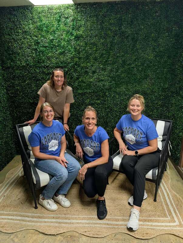 Four women in matching blue t-shirts sit in front of a green foliage wall, smiling together in a cozy setting.