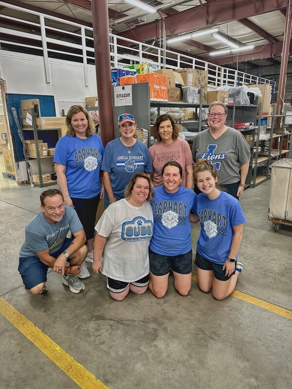 Eight people are posing and smiling inside a warehouse with shelves and boxes in the background. Most are wearing blue shirts with school or team logos.