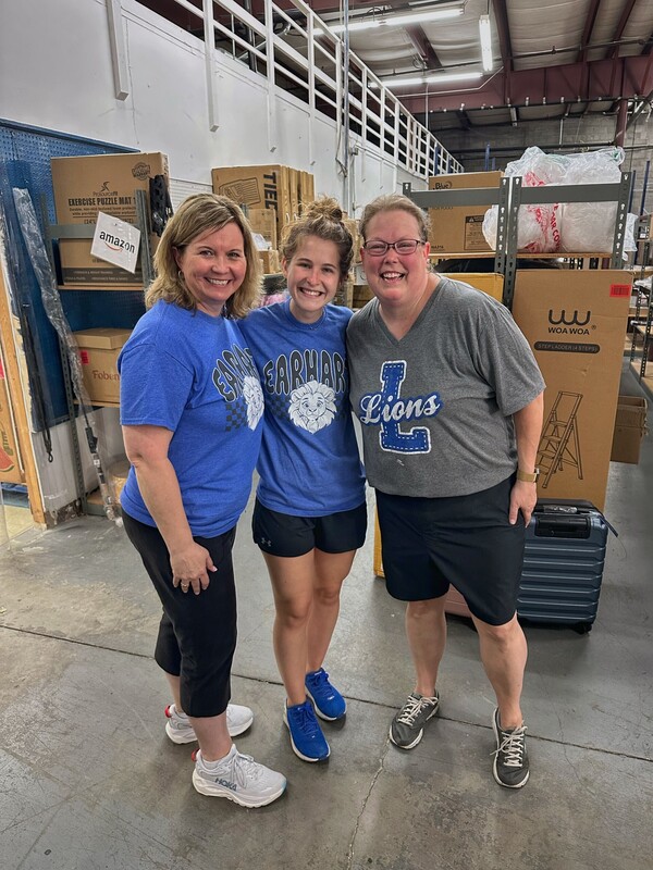 Three women wearing blue shirts stand together, smiling, in a warehouse setting with shelves, boxes, and equipment visible in the background.