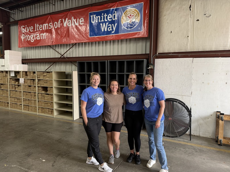 Four women smiling in a warehouse, standing in front of a red "United Way" banner and organized shelves of cardboard boxes.