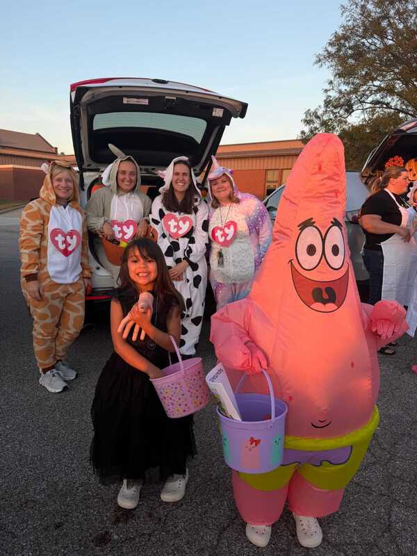 At a school Halloween event, women in colorful costumes pose beside a car with its trunk open, while a person in a bright Patrick Star suit waves.