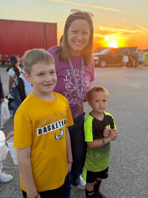 A smiling woman stands with two young boys in bright shirts at an outdoor event, with trucks behind them and a warm sunset glow.
