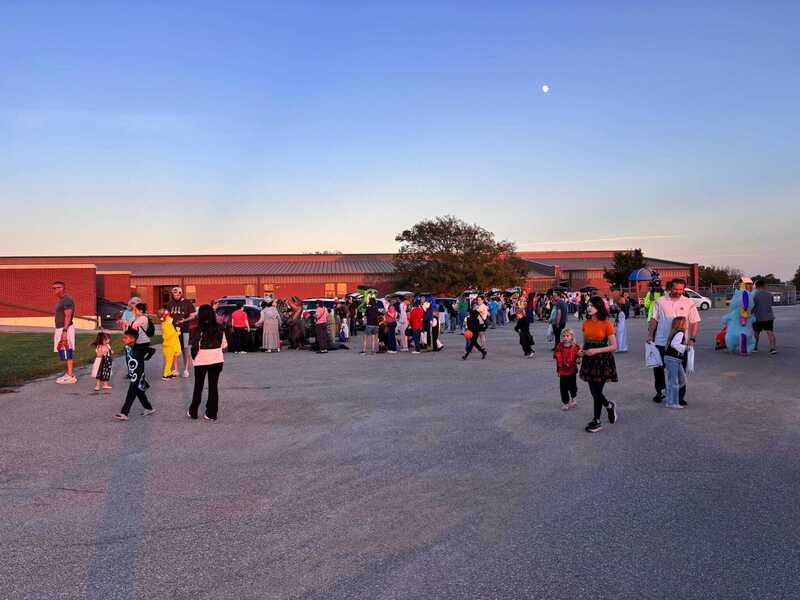 Families gather in costume beside parked cars in a school parking lot at sunset, creating a lively, welcoming scene for a community event.