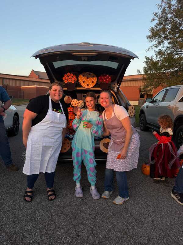 Three smiling women pose beside a car decorated with orange-and-black Halloween pumpkins, while a child in a red costume stands nearby in the parking lot.