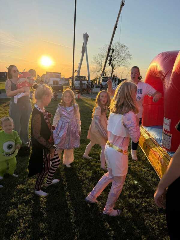 Children in costumes gather beside a red inflatable slide at a sunset fair, with an adult nearby; warm golden light creates a festive, joyful mood.